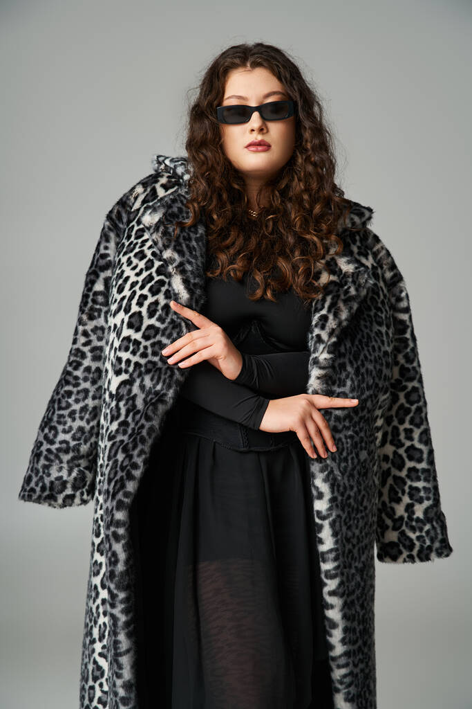mulher curvilínea confiante em óculos de sol de pé com casaco de pele de leopardo no fundo cinza - Foto, Imagem