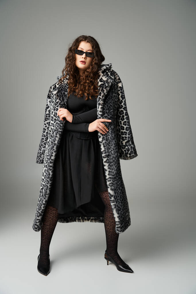 kurvige Frau in Sonnenbrille mit Leopardenfell-Mantel und weit auseinander stehenden Beinen auf grauem Hintergrund - Foto, Bild