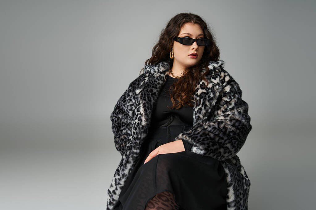 chica con curvas en abrigo de piel de leopardo y gafas de sol sentado en la silla con las piernas cruzadas y mirando a un lado - Foto, imagen