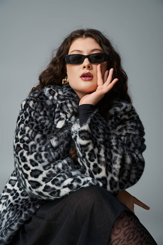 krásná zakřivená žena v leopard kožich kabát a sluneční brýle sedí na židli a odpočívá bradu na ruce - Fotografie, Obrázek