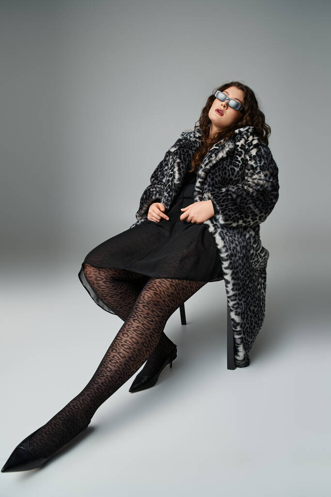 atrakcyjny plus rozmiar kobieta w lampart futro i okulary przeciwsłoneczne zrelaksowany siedzi na czarnym krześle - Zdjęcie, obraz