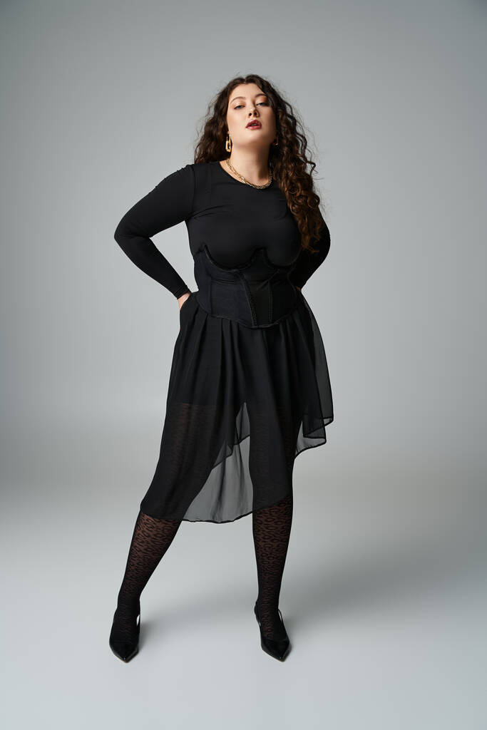 stylische Plus Size Frau im schwarzen Outfit mit den Händen auf dem Rücken vor grauem Hintergrund - Foto, Bild