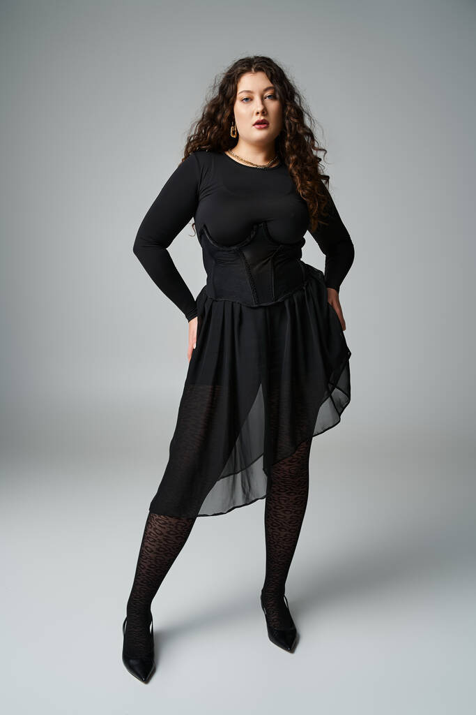 красива жінка плюс розмір в чорному вбранні з кучерявим волоссям і руками на стегнах на сірому фоні - Фото, зображення