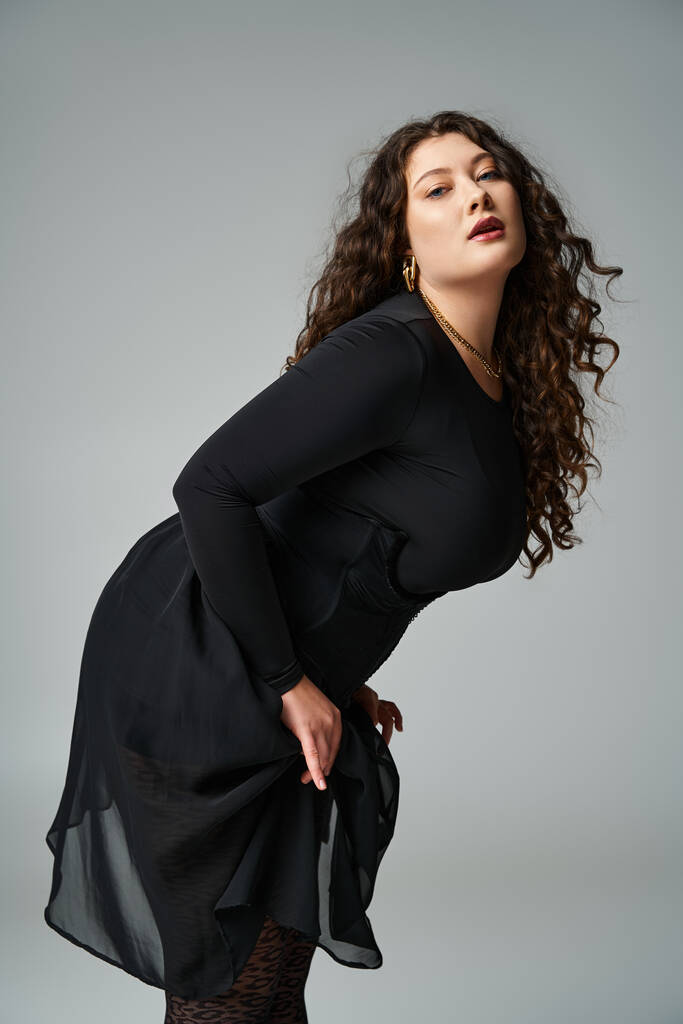 verleidelijk plus size vrouw in zwart outfit leunend naar voren zijwaarts tegen grijze achtergrond - Foto, afbeelding