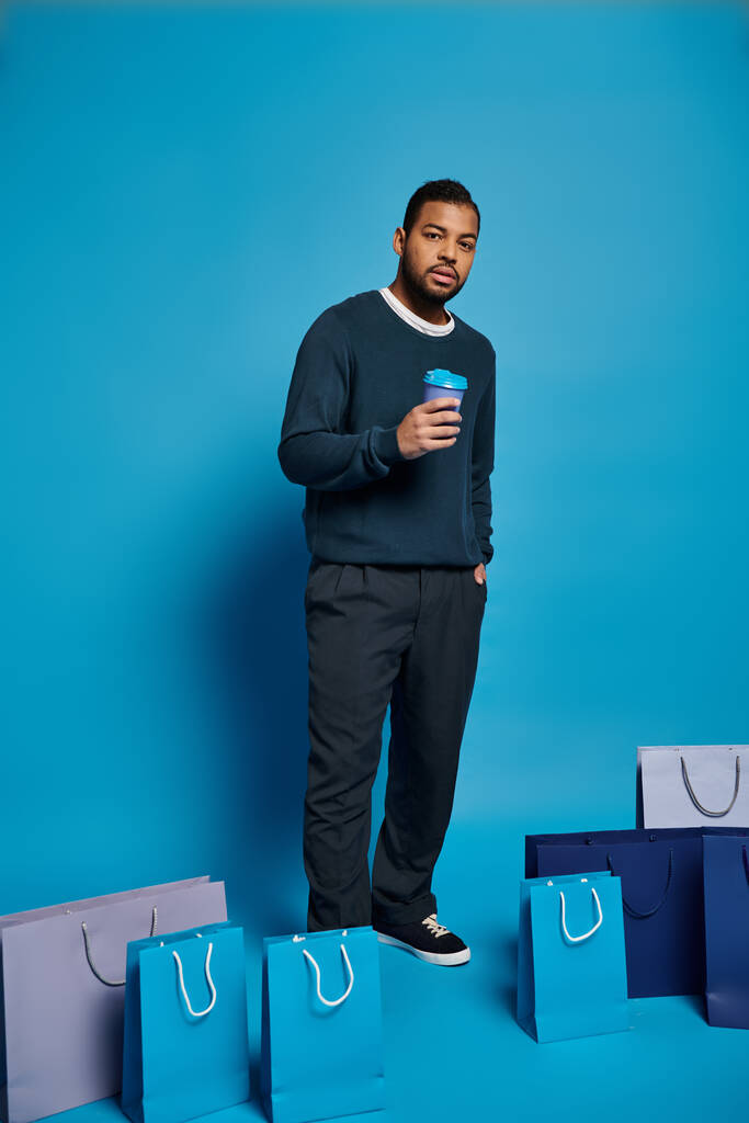 homem americano africano em azul escuro roupa em pé com sacos de compras, copo de papel e mão no bolso - Foto, Imagem