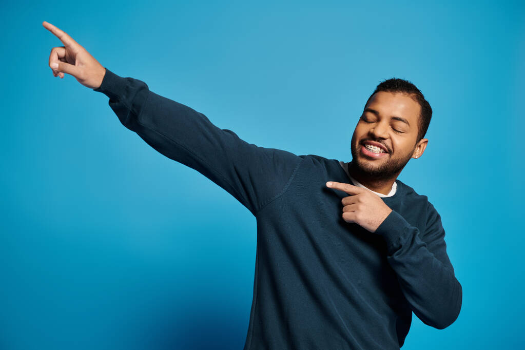 cheerful african american man in his 20s showing fingers to side with closed eyes on blue background - Photo, Image