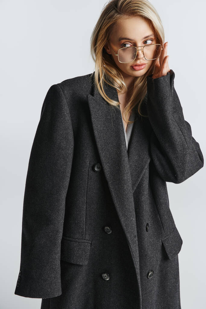 elegante mujer de moda con el pelo rubio y gafas elegantes en el abrigo negro posando y mirando hacia otro lado - Foto, imagen