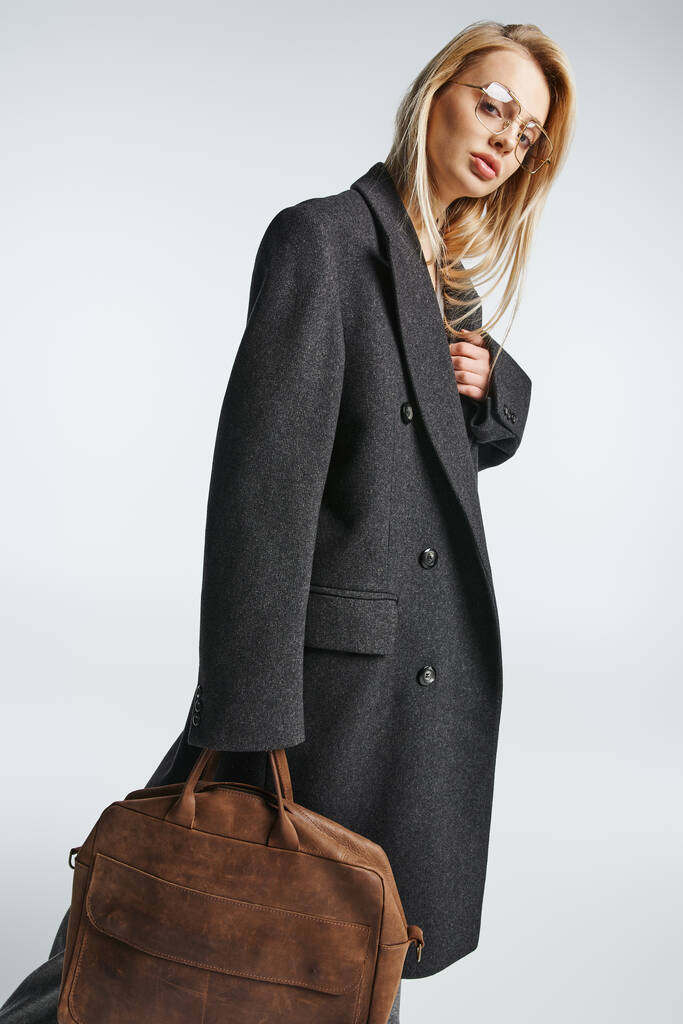 mooie modieuze vrouw met bril en bruine tas in zwarte elegante jas op zoek naar camera - Foto, afbeelding