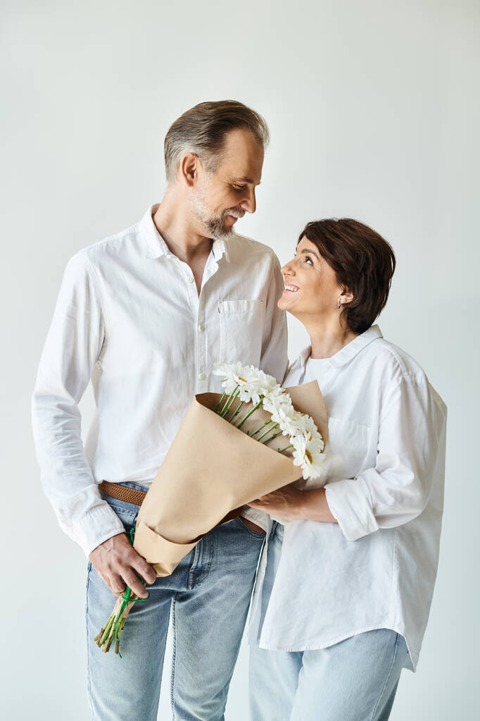 Beyaz gömlekli orta yaşlı bir adam gri arka planda çekici bir kadına çiçek sunuyor. - Fotoğraf, Görsel