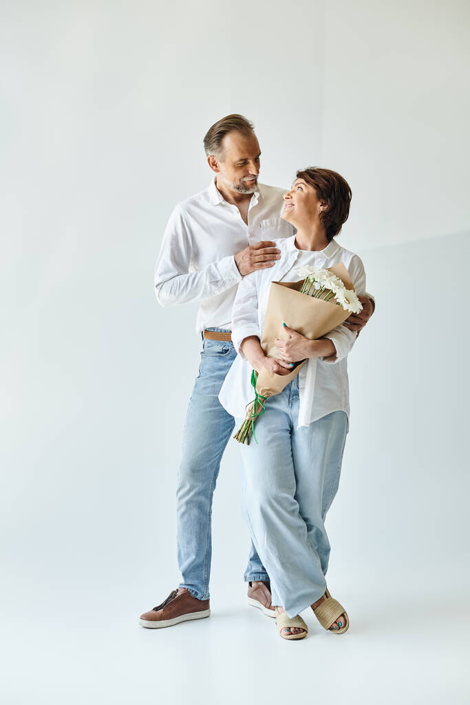 Full length shot of middle aged couple with flowers on grey background and looking at each other - Photo, Image