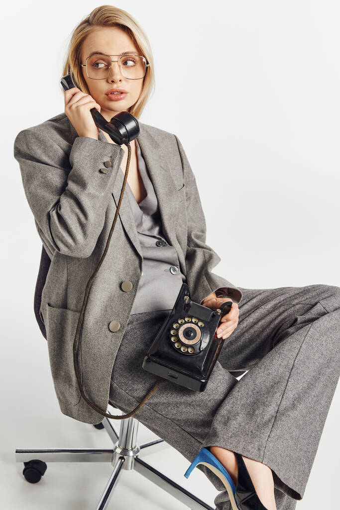 приваблива шикарна жінка зі стильними окулярами в сірому костюмі, сидячи на стільці і розмовляючи ретро телефоном - Фото, зображення