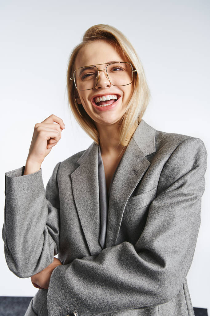 mujer atractiva alegre con gafas en traje elegante gris posando sobre fondo gris y mirando hacia otro lado - Foto, imagen
