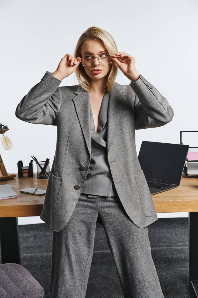 bela mulher de negócios elegante em terno inteligente cinza posando ao lado da mesa no escritório e olhando para longe - Foto, Imagem