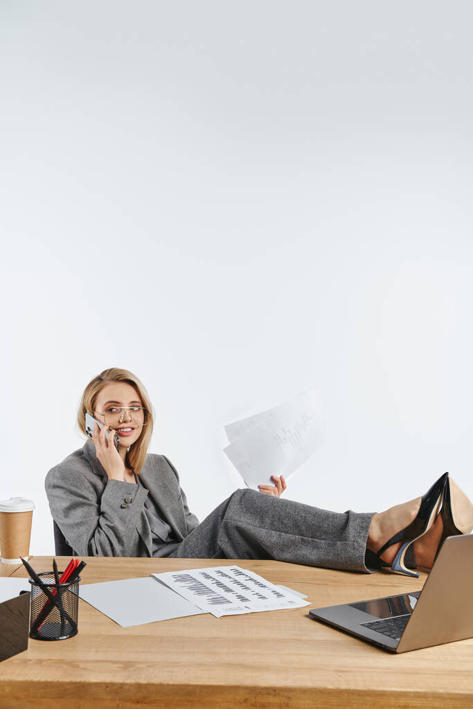 Радостная деловая женщина в сером костюме в стильных очках разговаривает по телефону, сидя за столом - Фото, изображение