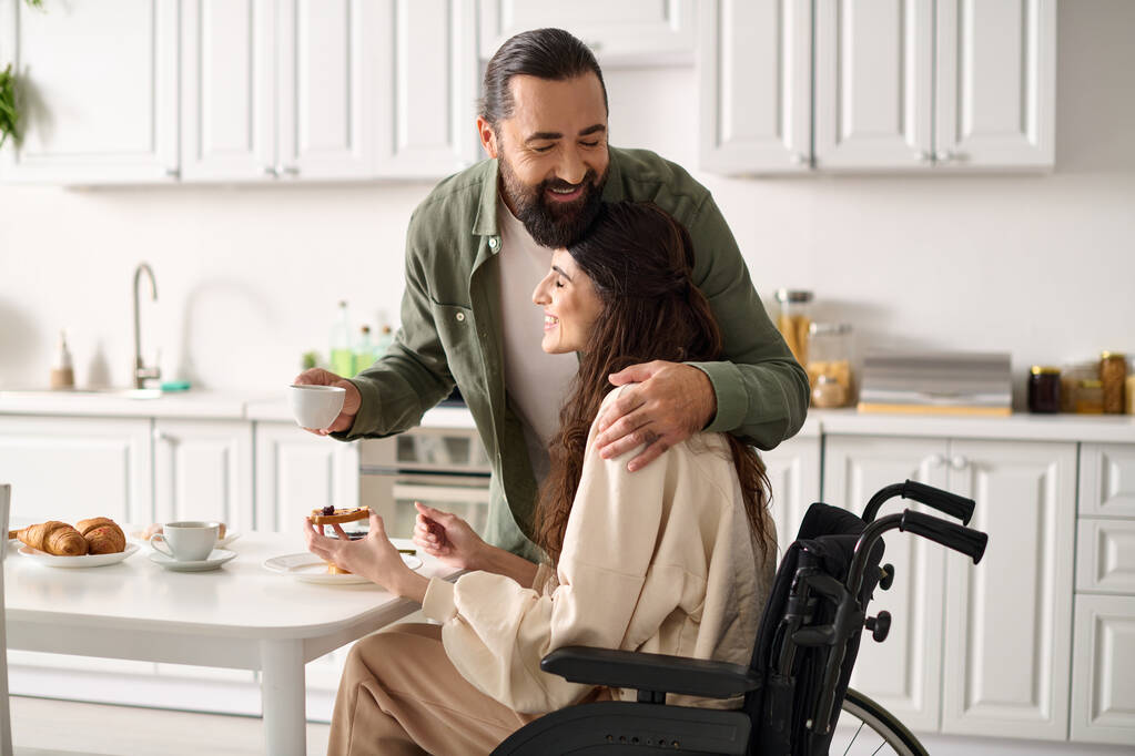 joyful attractive woman with disability in wheelchair eating breakfast with her loving husband - Photo, Image