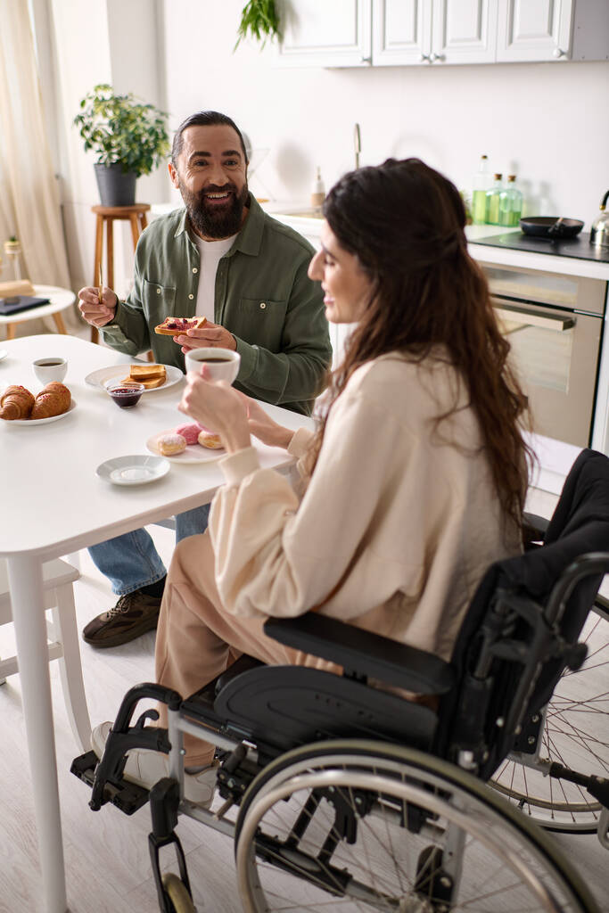 радостная женщина с включённостью в инвалидное кресло ест сладости за завтраком со своим красивым мужем - Фото, изображение