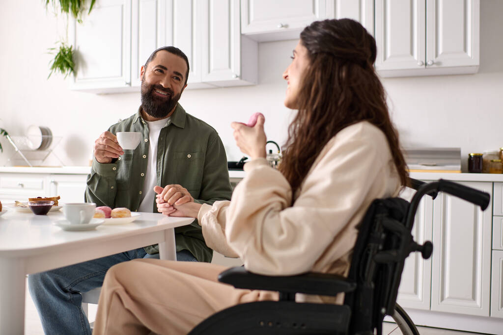 radostná žena s inkluzivitou v invalidním vozíku jíst sladkosti u snídaně se svým krásným manželem - Fotografie, Obrázek
