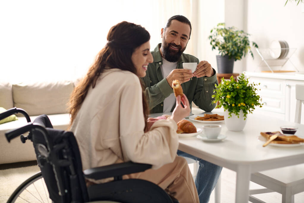 positive man having great time at breakfast with his beautiful disabled wife that eating croissant - Photo, Image