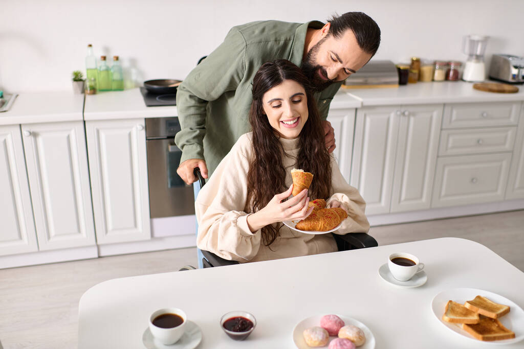 alegre hombre teniendo gran tiempo en el desayuno con su hermosa esposa discapacitada que comer croissant - Foto, Imagen