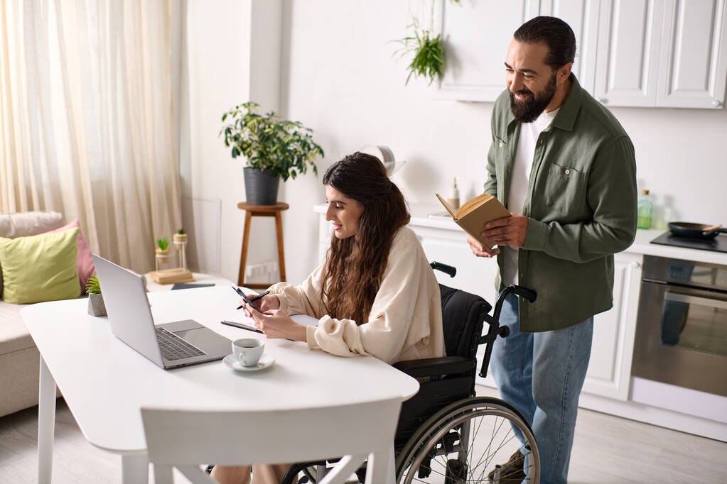θετική ανάπηρη γυναίκα σε αναπηρική καρέκλα που εργάζεται εξ αποστάσεως στο σπίτι δίπλα στον σύζυγο της που διαβάζει - Φωτογραφία, εικόνα