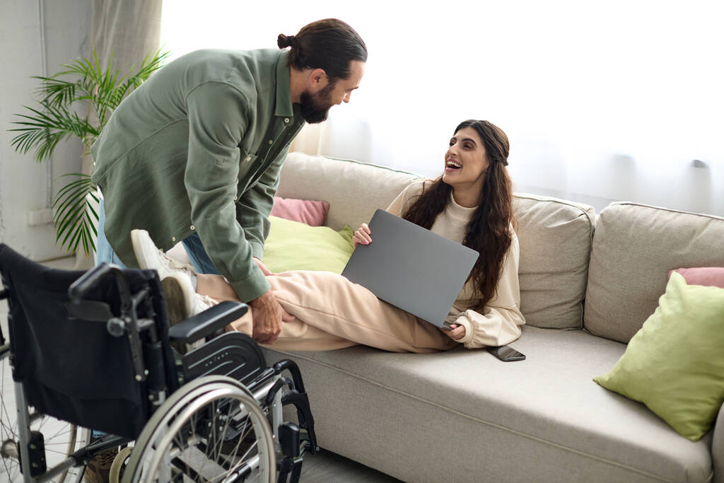 ελκυστική γυναίκα με αναπηρία κινητικότητας κάθεται στον καναπέ με φορητό υπολογιστή δίπλα στον αγαπημένο σύζυγό της - Φωτογραφία, εικόνα