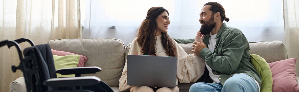 alegre hombre mirando a la computadora portátil al lado de su discapacitada hermosa esposa alegre mientras está sentado en el sofá en casa - Foto, imagen