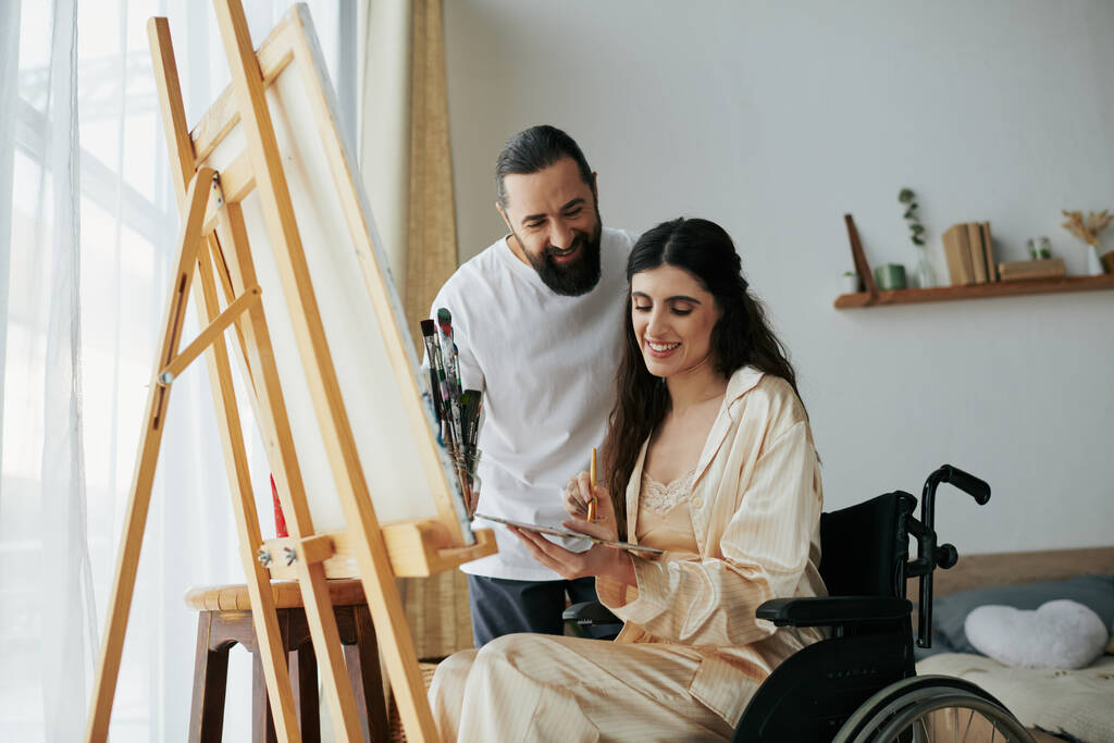atractivo barbudo hombre viendo su discapacitado hermosa esposa en silla de ruedas pintura en caballete en casa - Foto, imagen