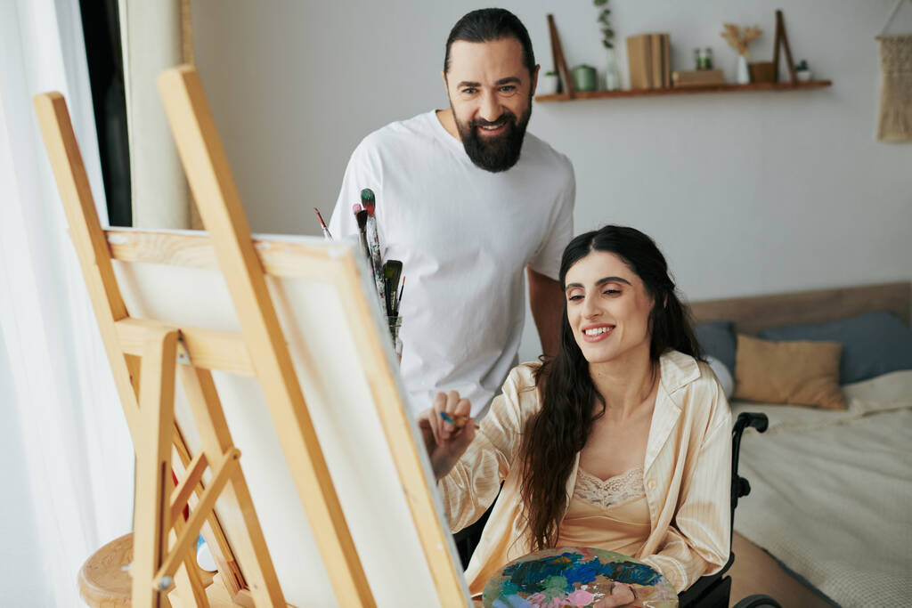 atractivo barbudo hombre viendo su discapacitado hermosa esposa en silla de ruedas pintura en caballete en casa - Foto, Imagen