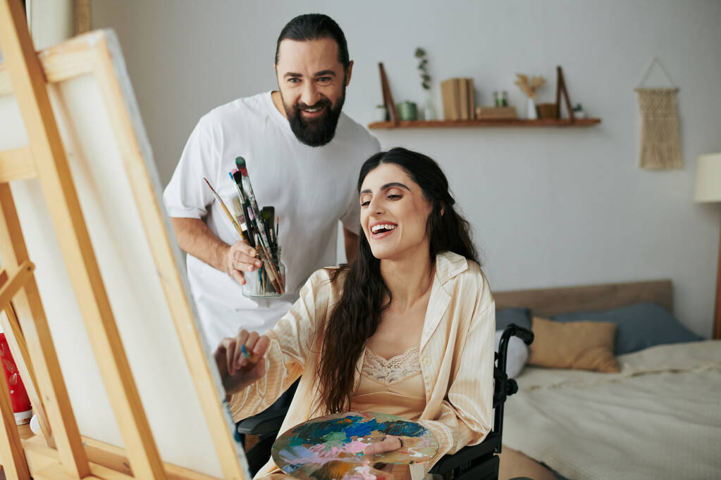 milující veselý manžel pomáhá jeho inkluzivní dobře vypadající manželka malovat na stojan, zatímco doma - Fotografie, Obrázek