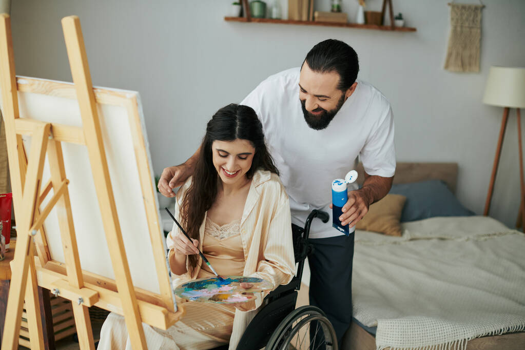 cuidado alegre pareja de barbudo hombre y discapacitado mujer pintura en caballete juntos en casa - Foto, imagen