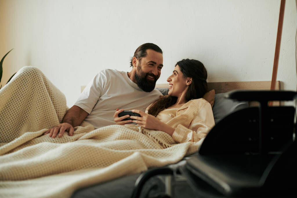 красивая радостная женщина с ограниченной подвижностью лежит в постели рядом со своим бородатым любящим мужем - Фото, изображение