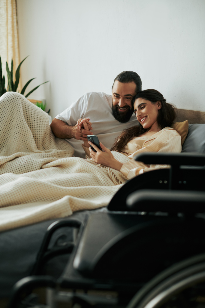 freudige Frau mit Gehbehinderung, die mit Handy neben ihrem bärtigen, liebevollen Ehemann im Bett liegt - Foto, Bild