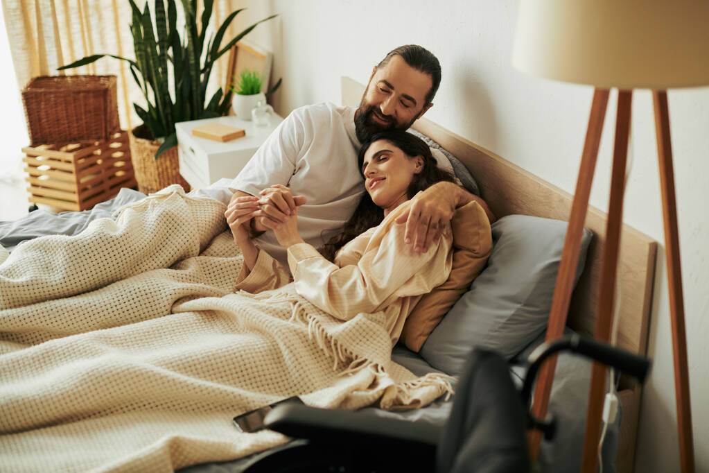 atractiva mujer alegre con discapacidad de movilidad tumbada en la cama junto a su marido barbudo amoroso - Foto, Imagen