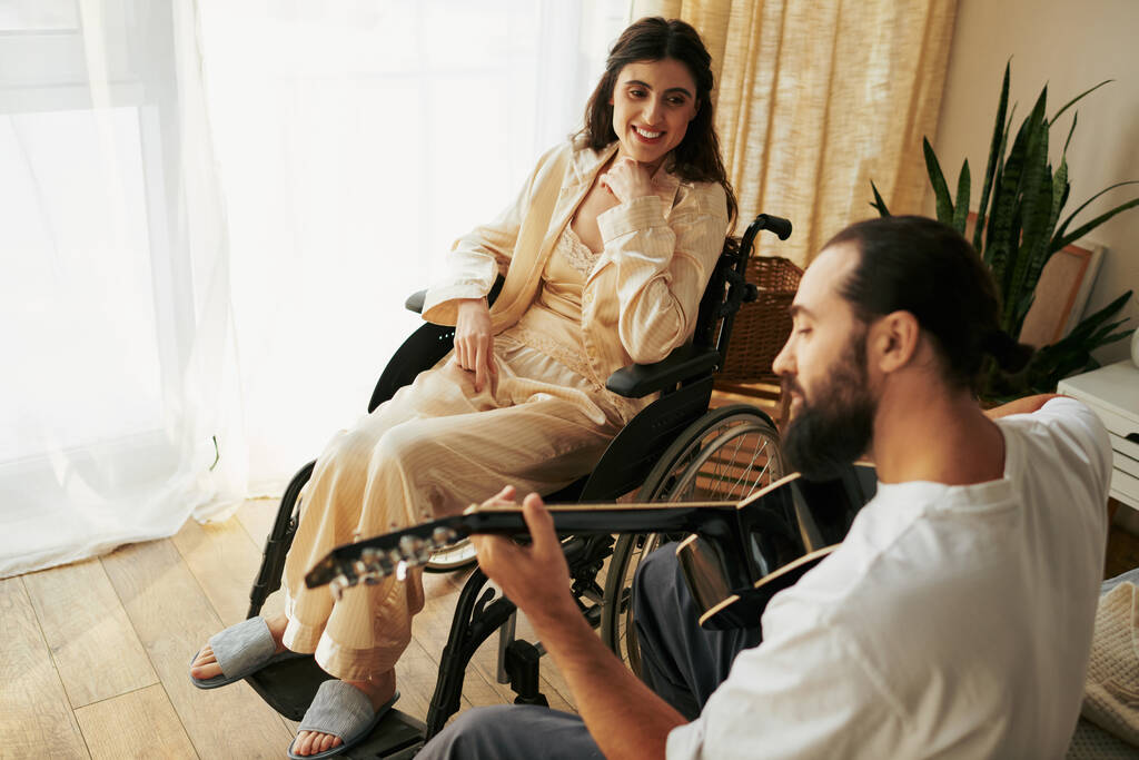 radosna kobieta z niepełnosprawnością na wózku inwalidzkim ogląda swojego brodatego męża grającego dla niej na gitarze - Zdjęcie, obraz