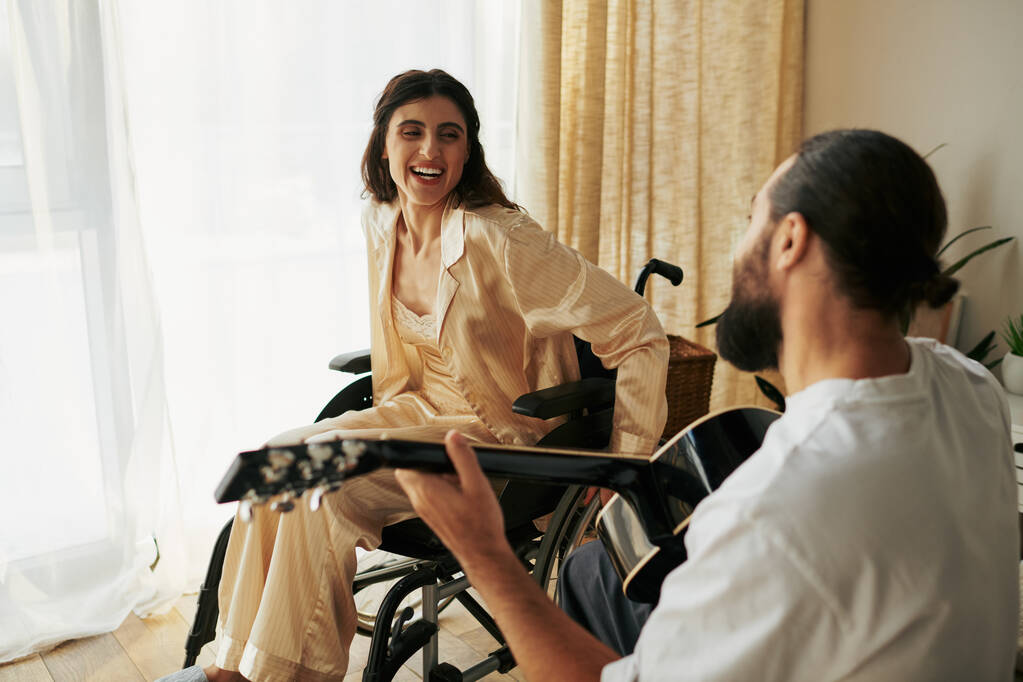 fröhliche Frau mit Behinderung im Rollstuhl, die ihrem bärtigen Mann dabei zusieht, wie er für sie Gitarre spielt - Foto, Bild