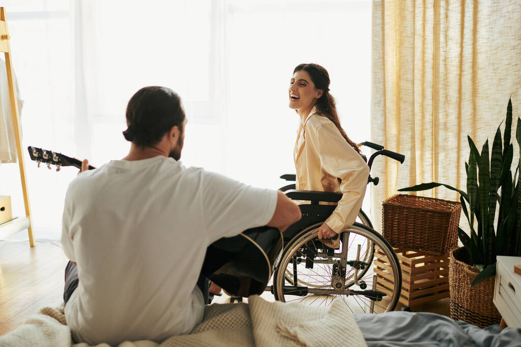 mujer alegre con discapacidad en silla de ruedas viendo a su marido barbudo tocar la guitarra para ella - Foto, Imagen