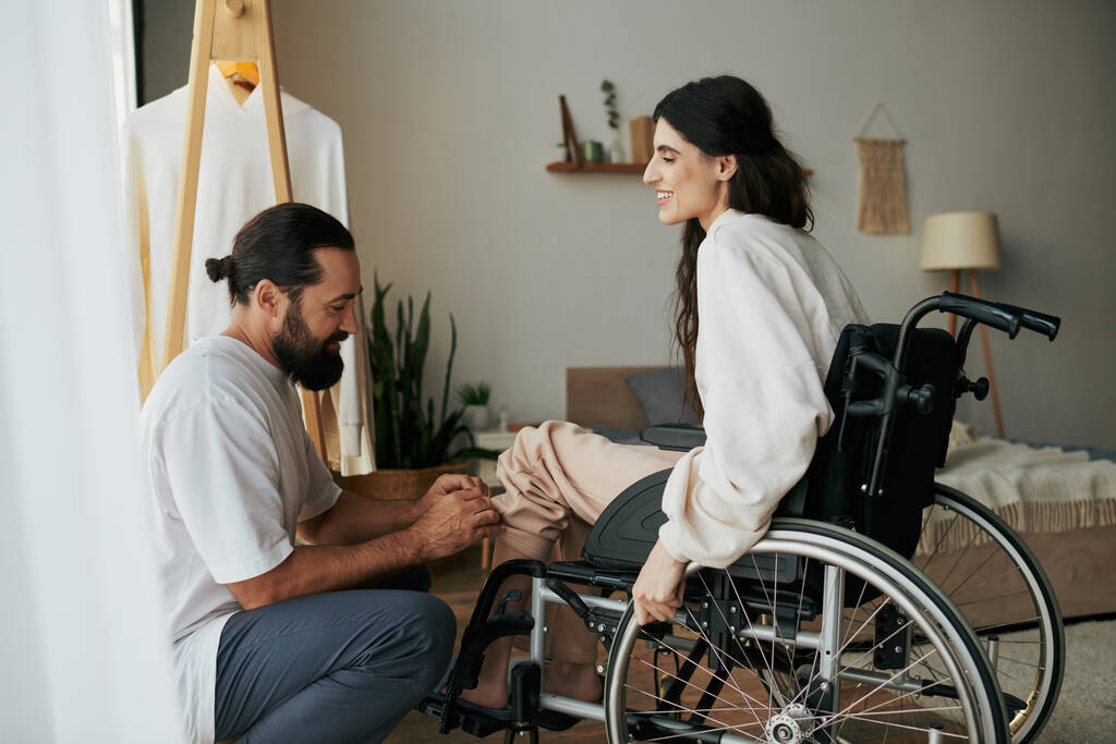 bearded loving man helping his inclusive wife on wheelchair to get dressed while in bedroom - Photo, Image