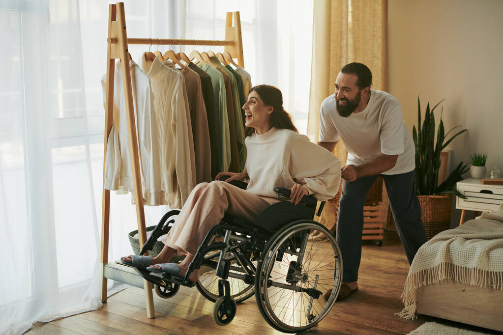 красивий дбайливий чоловік допомагає своїй інклюзивній дружині на інвалідному візку одягатися в спальні - Фото, зображення