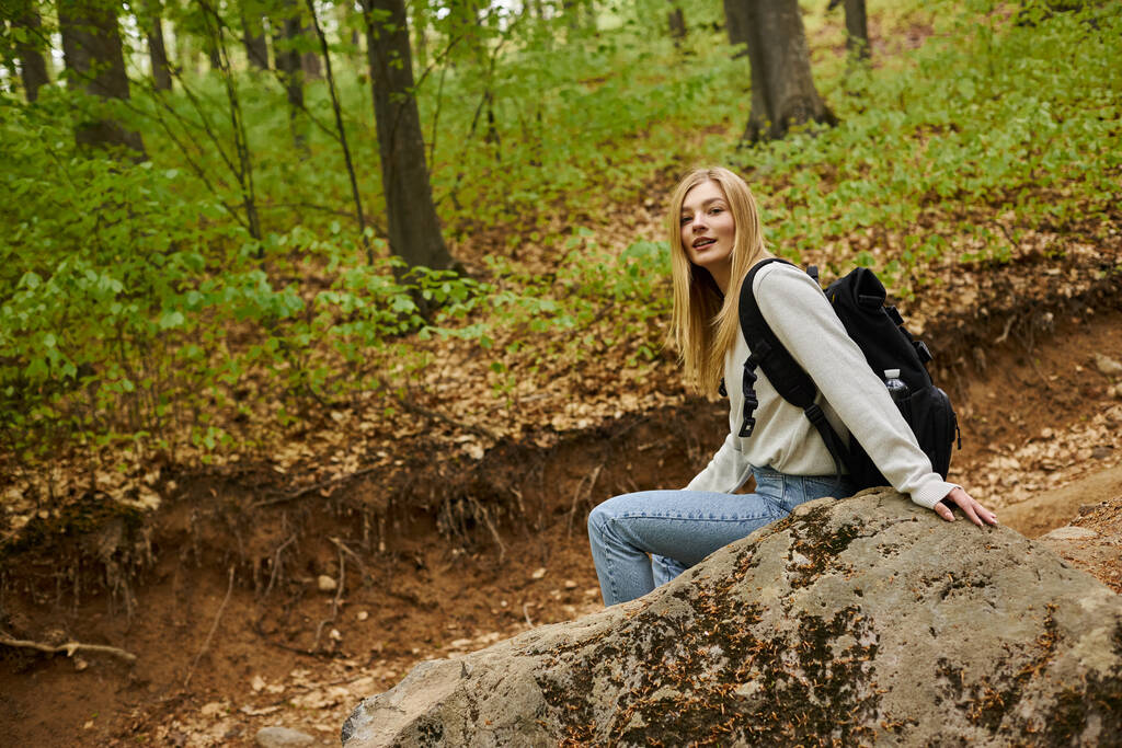 Χαμογελώντας χαλαρή ξανθιά πεζοπόρος φορώντας πουλόβερ και σακίδιο κάθεται σε ένα βράχο στο τοπίο του δάσους - Φωτογραφία, εικόνα