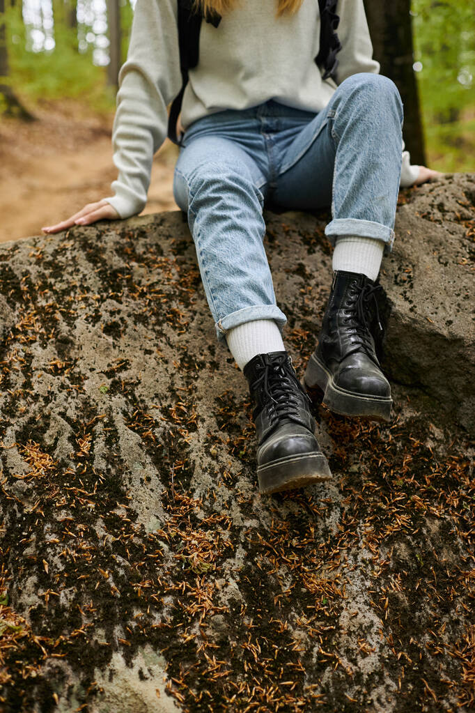 Cropped image of female hiker legs wearing jeans and hiking boots sitting on rock in forest - Photo, image