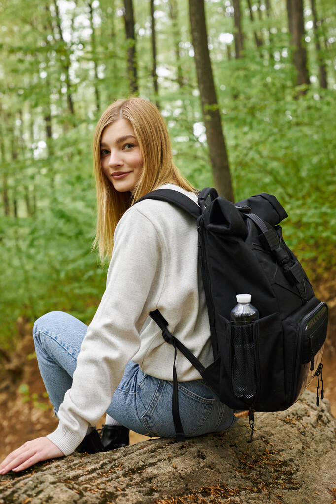 Achteraanzicht portret van zorgeloze blonde wandelaar in gezellig, trui zittend in bos en kijkend naar camera - Foto, afbeelding