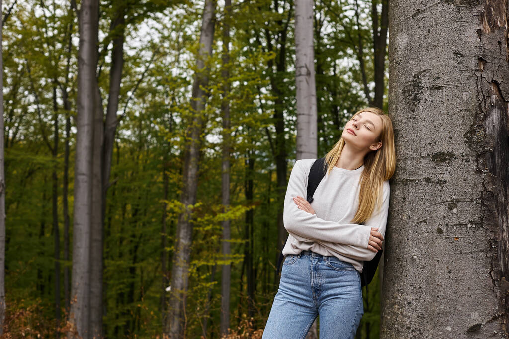 Portret zrelaksowanej blondynki turystki w lesie opierającej się o drzewo z zamkniętymi oczami - Zdjęcie, obraz