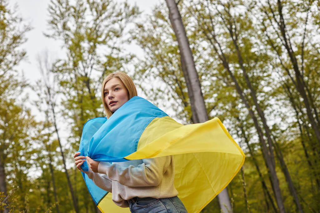 Beautiful patriotic blonde woman standing in the forest covering body with Ukrainian flag - Photo, Image