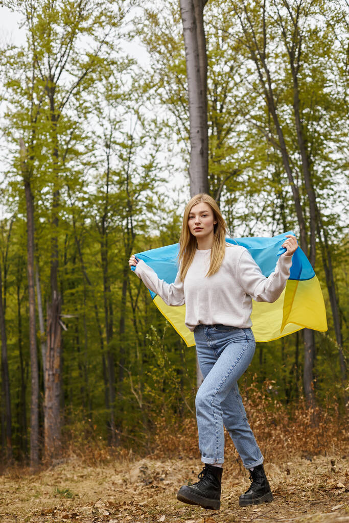 Όμορφη ξανθιά γυναίκα στέκεται στο δάσος καλύπτει το σώμα με την ουκρανική εθνική σημαία ως ακρωτήριο - Φωτογραφία, εικόνα