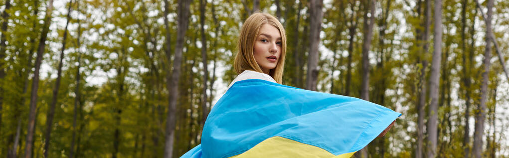 Back view of female young blonde hiker, holding the Ukrainian flag and turning to camera, banner - Photo, Image