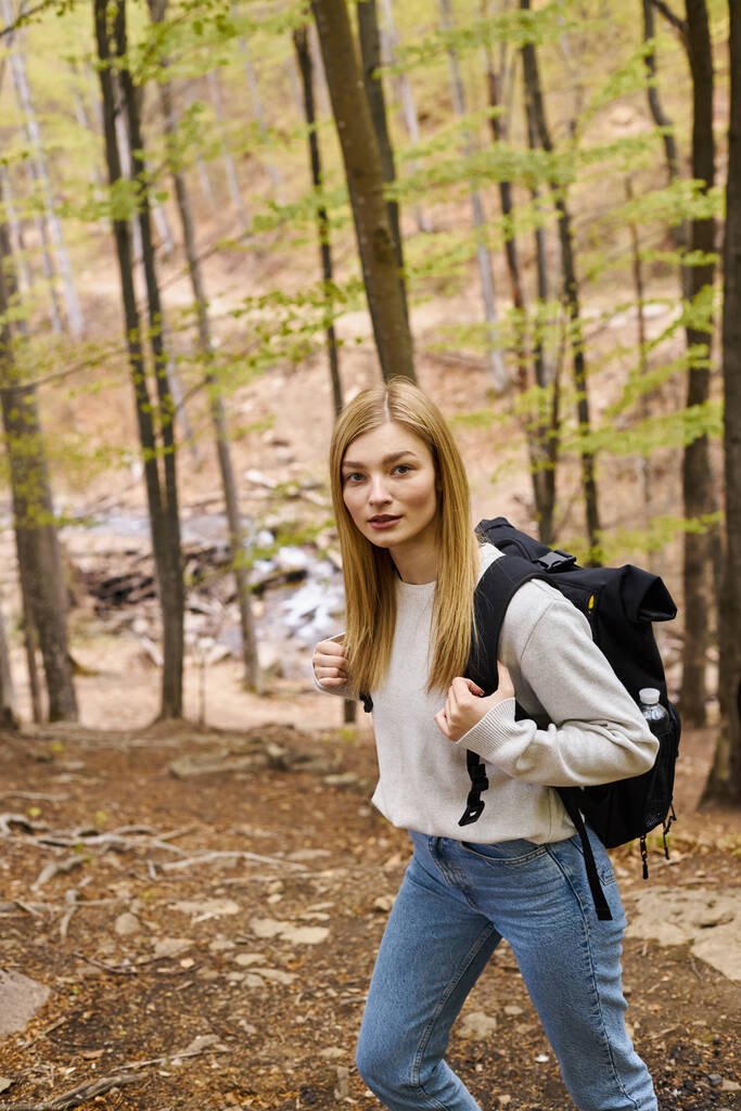 Neugierige blonde Wanderin mit Reiserucksack schlängelt sich durch den Wald und blickt in die Kamera - Foto, Bild