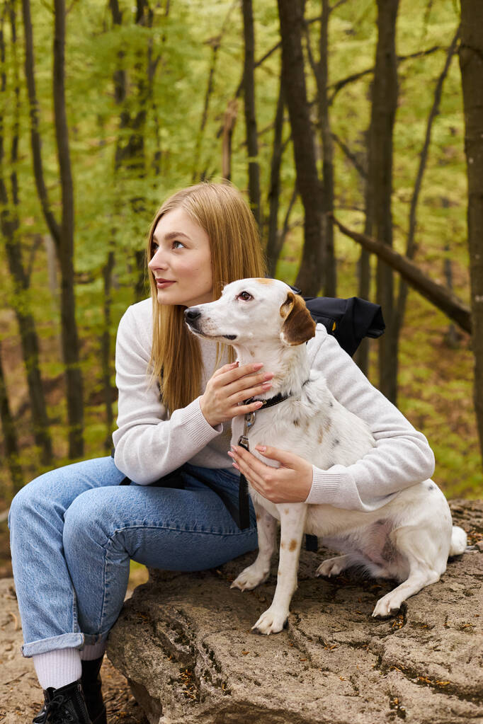 Smiling blonde woman hugging her dog, while having a halt on forest trip, both looking away - Photo, Image