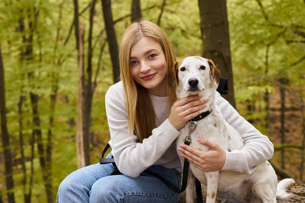 Uśmiechnięta blondynka przytula psa, zatrzymując się na leśnej wycieczce, patrząc w kamerę - Zdjęcie, obraz