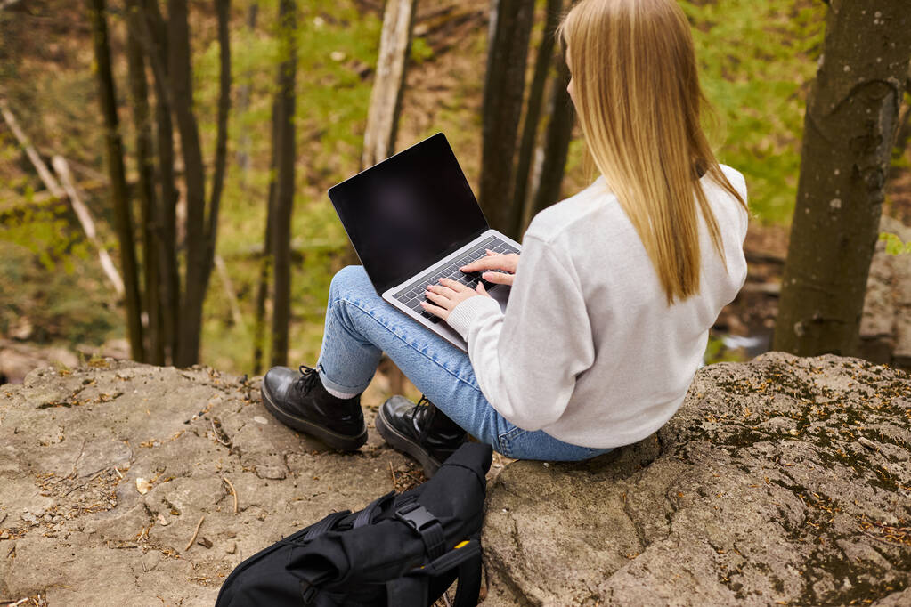 Back view of blonde female hiker sitting on boulder in forest with laptop on her knees with backpack - Photo, Image