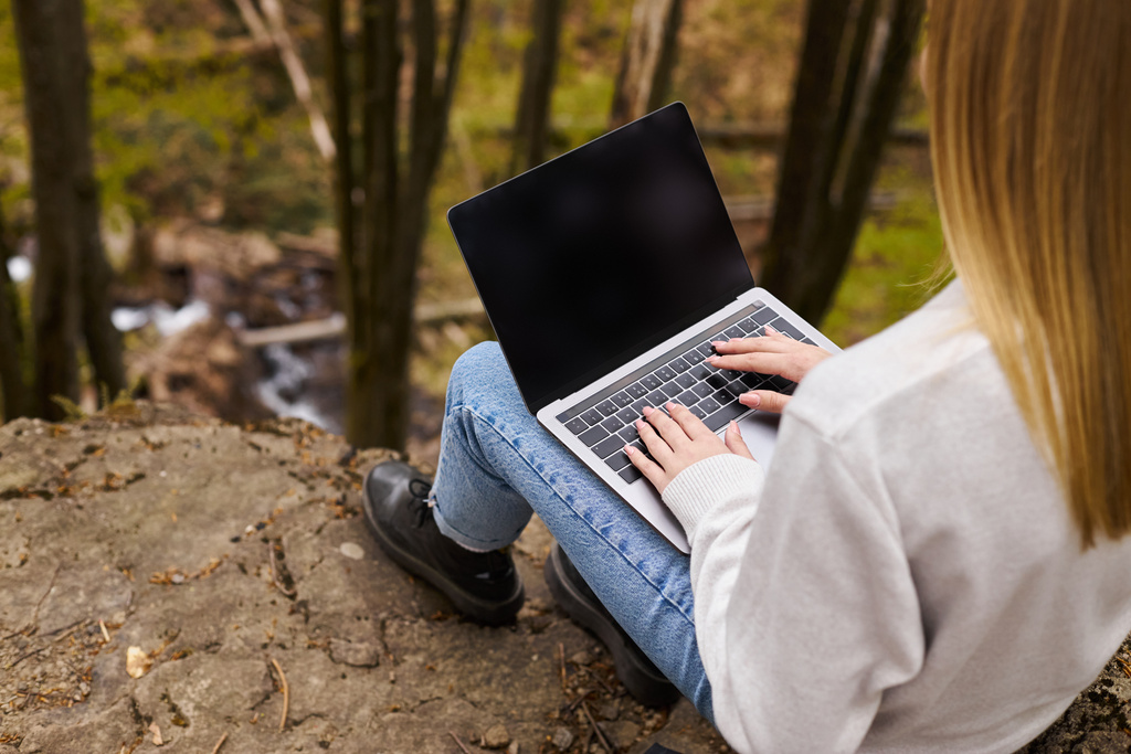 Πίσω όψη μιας ξανθιάς γυναίκας που κάθεται σε ένα μεγάλο βράχο στο δάσος, χρησιμοποιώντας ένα φορητό υπολογιστή για απομακρυσμένη εργασία - Φωτογραφία, εικόνα